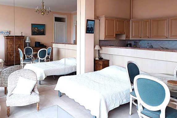 Chambre lumineuse avec deux lits, mobilier en bois clair et cuisine ouverte.