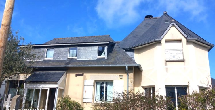 Maison à deux étages avec toit en ardoise, fenêtres blanches et jardin. Ciel bleu.